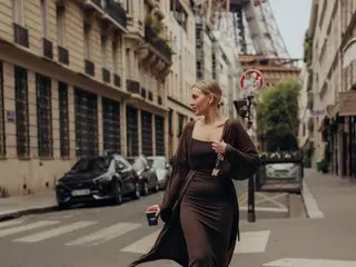 AurorraAksness poses on a city street wearing a dark, low-cut dress that shows cleavage, with her blonde hair styled down.