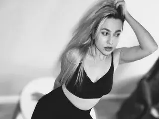 AvaBlueX poses with her hands in her long blonde hair, wearing a black bra and black bottoms against a light background.