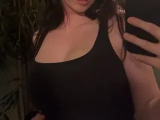 Avallanchee poses in a black tank top, showing her long dark hair and cleavage in a dimly lit setting.