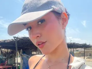 Azulhorny poses close-up, wearing a grey baseball cap and a black strap, showing her face and shoulders against a bright outdoor background.