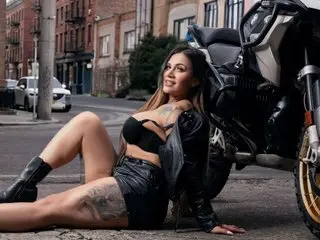 AzunaStar poses in a black leather outfit, showing off her long hair and tattoos while sitting outdoors on a city street.