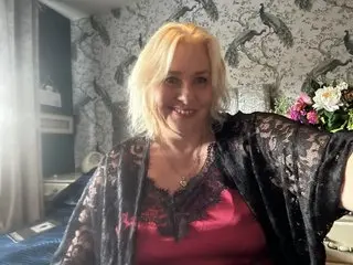 BBWQueen69 smiles while wearing a red lace top and a black lace open robe, showing her blonde hair and cleavage in a room with floral wallpaper.