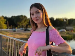 BabLumii poses outdoors in a pink top, showing her long brown hair and shoulders against a soft sunset background.