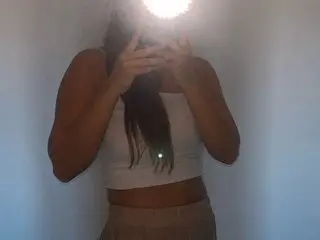 BabeAngelFace poses in a tan crop top, holding her hands up near her face against a plain white wall.