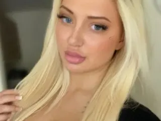 Blonde woman with glamorous makeup and long hair posing in a soft-lit indoor setting.
