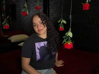 Baddie78Kamila poses in a black t-shirt with a purple graphic, showing her long dark curly hair against a dark background.