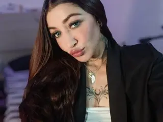 Bahianabelly poses with long dark hair, wearing a black blazer over a white top, showing a large black ink tattoo on her chest.
