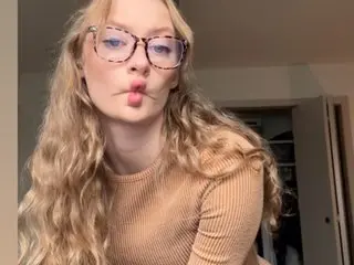 Bailey_AnnXXX with long blonde wavy hair, wearing a tan ribbed long-sleeve top and glasses, looking at the camera with a pouting expression.