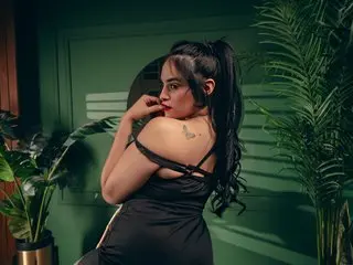 BarbaraSaen poses with her long dark hair and bare shoulders, wearing a black strap top against a dark green wall.