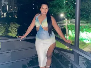 Barbara_Lopez poses outdoors at night wearing a light blue cut-out bodysuit that shows her cleavage and midriff, standing by a balcony railing.