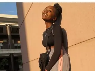 Smiling young Black woman in a black long-sleeve top and grey skirt standing against a sunlit wall.