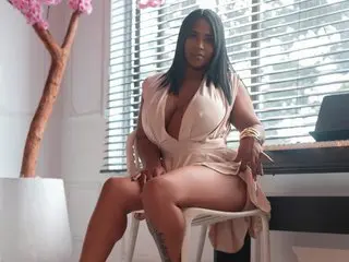 BeckyVega poses in a low-cut beige dress, showing cleavage and her legs while sitting in front of white blinds.