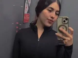BellaLuuna poses for a mirror selfie wearing a black zip-up jacket, showing her dark hair and face against a grey wall.