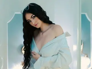 BellaMillers poses with long dark hair and a deep cleavage, wearing a white off-the-shoulder shirt against a light blue background.