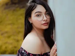 BellaSophiee poses with long dark hair, wearing a black floral top, looking over her shoulder against a soft outdoor background.