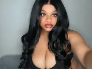 BellaWella18 poses with long, wavy black hair, wearing a black top that shows off her cleavage against a plain background.