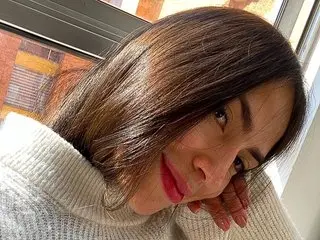 Bellasmmith smiles while resting her chin on her hand, showing her long brown hair and white turtleneck sweater.