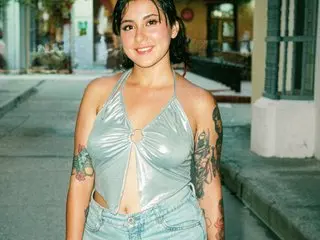 Bellauwu poses outdoors wearing a silver metallic halter top and light blue denim jeans, showing her shoulders and cleavage.