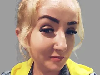Close-up portrait of a blonde woman in a yellow hoodie with a neutral expression against a gray background.