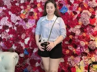 Benna69 — Smiling young woman in a light blue blouse and black shorts standing before a vibrant pink floral wall.