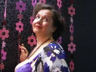 BestMILF smiles while wearing a purple lace top and a floral patterned blouse against a black background with pink flower decorations.