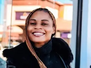 Smiling woman with long blonde braids wearing a black fuzzy coat in a bright outdoor urban setting.