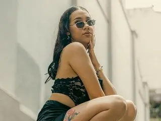 BibiSugarx poses outdoors with long dark hair, wearing a black lace crop top and black shorts, showing off her bare shoulders and legs.