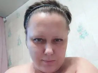 BigAss111 looks directly at the camera with her hair pulled back, showing her bare shoulders and face against a light patterned wall.