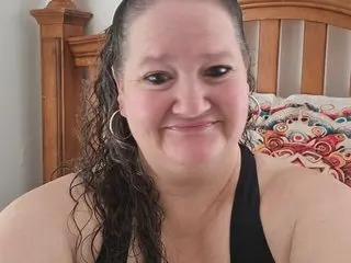 Smiling woman with long curly hair wearing a black halter top in a bedroom setting.