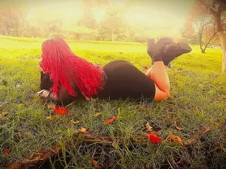 Bigassnolimits66 has long red hair and is lying on her stomach in a grassy field, wearing black bottoms that show her bare legs and buttocks.