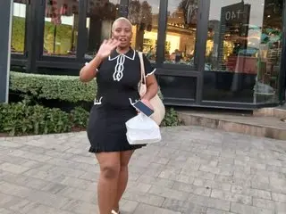 Bigbootycurvemilf stands outside wearing a black short-sleeved dress with white trim, holding a white handbag and posing with one hand near her face.