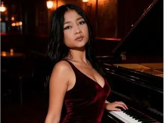 BiiancaShermann poses with long dark hair, wearing a low-cut red velvet tank top while sitting next to a black piano.