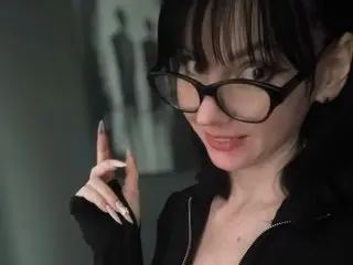 BleckMamba poses with a smirk, wearing black glasses and a black zip-up top with her dark hair styled in a fringe.
