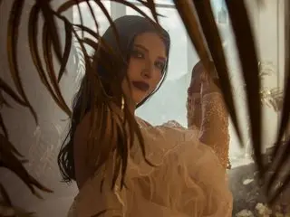 BlissAura poses with long dark hair and dark lipstick, wearing a white ruffled top while framed by tropical palm leaves.