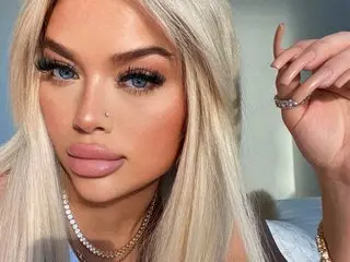 Glamorous blonde woman with blue eyes and long lashes wearing gold jewelry in soft natural lighting.
