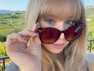BlondieBliss poses close-up, wearing dark sunglasses and showing her blonde hair and light eyes in a bright outdoor setting.