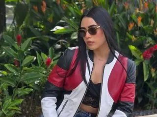 BlueCher poses outdoors wearing a black and red zip-up jacket over a black top, with her long dark hair down.