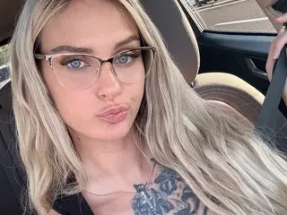 BonnieBazz poses with her long blonde hair down, wearing glasses and showing a hint of a tattoo on her chest.