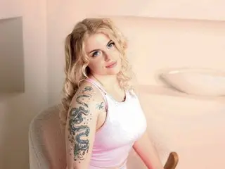 Bonny_Brok poses with long blonde hair, wearing a white tank top that shows off her tattoos on her arm and shoulder.