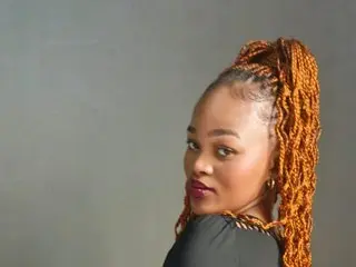 BootyCataleya looks over her shoulder with her hair styled in long, blonde-tipped braids against a plain grey background.