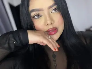 Brhiana_William poses with her hands near her face, showing her long dark hair and red lipstick against a plain background.