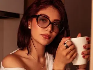 Young woman with glasses in a white top holding a coffee mug in a cozy indoor setting.