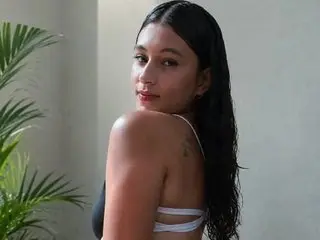 BriannaDuvvalle poses with her long dark hair, looking over her shoulder while wearing a white strappy top against a plain background.