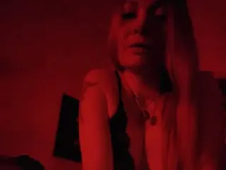 Blonde woman in a black lace top posing under moody red lighting in a dark room.