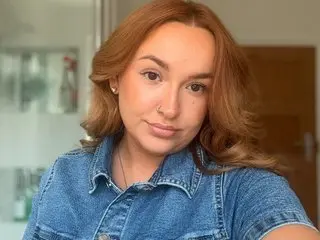 BritishMissPoppy poses for a close-up selfie, showing her face and shoulders while wearing a blue denim shirt and letting her long, wavy light-brown hair fall down.