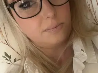 British_babe_jade looks at the camera with blonde hair and glasses, wearing a white floral print top.