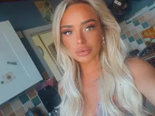 Britishblondeemily96 poses close-up, showing her blonde hair and face while wearing a light-colored top in a room setting.