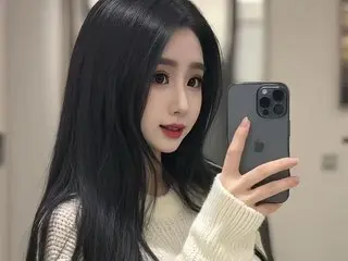 BritneyEvaa poses for a selfie with her long black hair down, wearing a white knit sweater against a neutral indoor background.