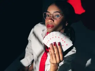 BrizzaHolguin poses with a hand near her face, wearing glasses and a white button-down shirt against a dark background.