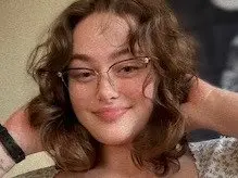 Brookiie_skrr smiles while posing with her hands behind her head, showing her wavy brown hair and light makeup.